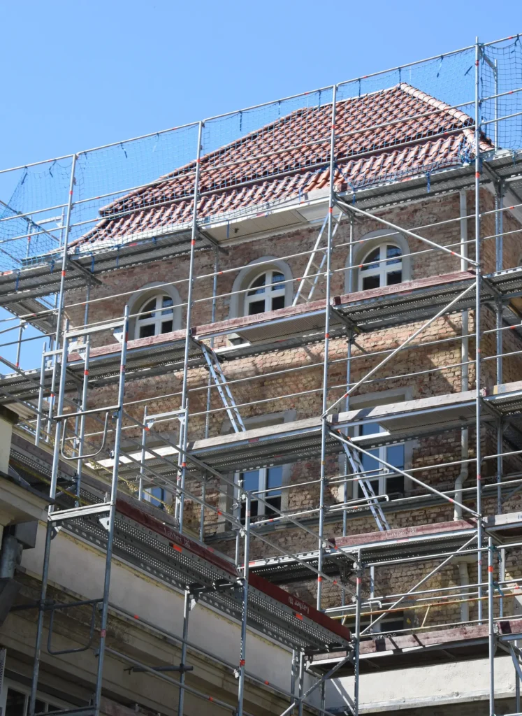 Échafaudage et rénovation d'un bâtiment ancien en briques Rénovation d'une façade en briques avec fenêtres cintrées, couverte par un grand échafaudage Layher sécurisé sous un ciel bleu.