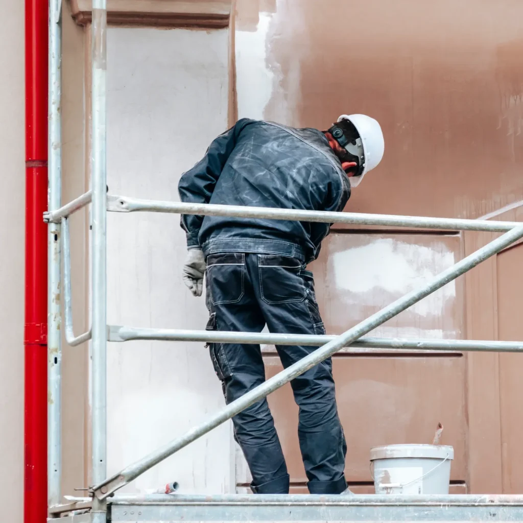 Rénovation de façade : ouvrier professionnel sur échafaudage Ouvrier sur échafaudage appliquant un enduit marron sur façade. Il porte un casque de sécurité blanc et des gants de travail.